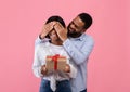 Happy black woman holding Valentine`s gift box, loving boyfriend covering her eyes on pink studio background