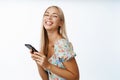 Happy blond modern girl, using mobile phone, smiling at camera, standing over white background. Cellular technology