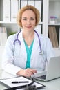 Happy blonde female doctor sitting at the table and typing by laptop computer. Medicine, healthcare and help concept Royalty Free Stock Photo