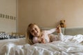 Cute blonde little girl in pajamas lying in white bed with teddy bear, awaking early in the morning before going to kindergarten. Royalty Free Stock Photo