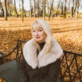 Happy blonde mature woman is sitting on bench in autumn park and thinking. Beautiful woman is relaxing in nature on sunny day. Royalty Free Stock Photo