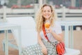 Happy blonde woman with curly light hair, holds fresh orange cocktail, being in high spirit, dressed casually, poses outdoor on be