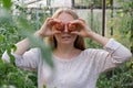 Happy blonde woman holding red tomatoes over eyes in hothouse. Female farmer having fun in green garden while harvesting Royalty Free Stock Photo