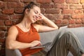 Happy blonde woman with laptop sitting on couch against brick wall. Remote work