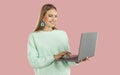 Happy blondy woman in mint sweatshirt doing remote job working on laptop on pink background.