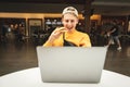 Happy boy with a burger in his hand, looking at the laptop screen and smiling, sitting at the table against the backdrop of a fast Royalty Free Stock Photo