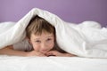 Happy boy hiding in bed under a white blanket or coverlet