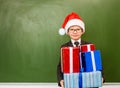 Happy boy in red christmas hat with gift boxes standing near empty green blackboard Royalty Free Stock Photo
