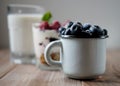 A small mug with blueberries, a glass of milk and granola with raspberries. Wooden background.
