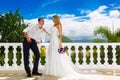 Happy bride and groom standing next to the stone gazebo amid beautiful tropical landscape. Sea, sky, flowering plants and palm tr Royalty Free Stock Photo