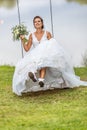 Happy bride in white wedding dress and flower bouquet in her hand swings outdoors next to a lake Royalty Free Stock Photo
