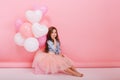 Happy brightful image of cute joyful little girl in tulle skirt sitting on present with balloons on pink