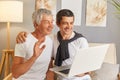 Happy brunette man embrace elderly father relaxing together in living room browsing internet on laptop family weekend time with