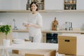 Happy brunette woman in casual outfit, moves into new house, unpackes boxes with kitchen dishes, holds plates, stands near table,