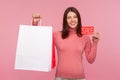 Happy brunette woman in pink sweater holding sale card and paper shopping bags, looking at camera with toothy smile, satisfied