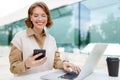 Happy business lady looks at phone screen with smile while sitting outside with laptop and coffee Royalty Free Stock Photo