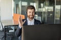 Happy businessman in headset working on computer and waving his hand to webcamera, having video call in office interior Royalty Free Stock Photo