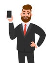 Happy businessman holding/showing smartphone in hand and holding hand on hip while standing against white background.