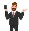 Happy businessman showing or holding smart phone, mobile, cell phone in hand and gesturing hand to copy space side away.