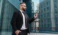 Happy businessman in suit talking on smartphone in wireless earphones standing near glass office building outdoors