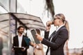 Happy businesswoman holding digital tablet and coffee to go outside of modern building, businesspeople in background Royalty Free Stock Photo