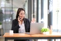 Happy businesswoman sitting at desk behind her laptop and talking with somebody on her mobile phone while working at Royalty Free Stock Photo