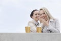 Happy businesswomen with disposable coffee cups sharing secrets against clear sky