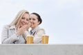 Happy businesswomen with disposable coffee cups sharing secrets against clear sky