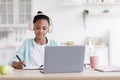 Happy busy teen black girl in earphones studying, doing homework at table with laptop in kitchen interior Royalty Free Stock Photo