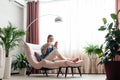 Happy calm Caucasian woman relax sit in comfort chair in cozy home interior use smartphone. Smiling young woman use