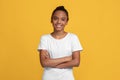 Happy calm teen black girl pupil in white t-shirt with crossed arms, isolated on yellow background