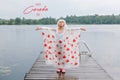 Happy Canada Day. Holiday card with text. Girl child in rain poncho with red maple leaves standing on wooden lake dock. Kid