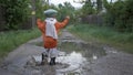 Happy carefree male kid in a hat and rubber boots have fun playing with leaves in a puddle on road