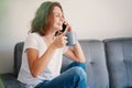 Happy casual beautiful woman is talking on a phone sitting on a sofa at home with cup of coffee Royalty Free Stock Photo