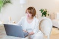 Happy casual beautiful woman working on a laptop sitting on the chair in the stylish white living room Royalty Free Stock Photo
