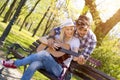 Happy caucasian couple sitting on a park bench, with him trying to teach her how to play guitar Royalty Free Stock Photo