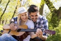 Happy caucasian couple sitting on a park bench, with him trying to teach her how to play guitar Royalty Free Stock Photo