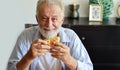 Happy caucasian elderly eating hamburger in living room with smiling face