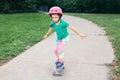 Happy Caucasian girl in pink helmet riding skateboard on road in park on summer day. Seasonal outdoors children activity sport. Royalty Free Stock Photo