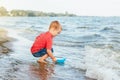Happy Caucasian Red-haired Toddler Child Boy Putting Blue Paper Boat In Water On Lake Sea Ocean