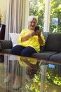 Happy caucasian senior woman sitting on sofa, using smartphone and smiling in sunny living room Royalty Free Stock Photo