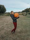 Happy Caucasian woman in jacket and red pants on farm carrying holding huge giant orange pumpkin