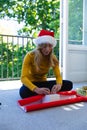 Happy caucasian woman in santa hat sitting on floor and packing gift at christmas at home