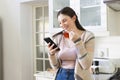 Happy caucasian woman standing, holding tea cup, using smartphone in kitchen at home, copy space Royalty Free Stock Photo