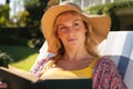 Happy caucasian woman wearing sunhat relaxing in sunny garden reading book, looking away, smiling Royalty Free Stock Photo