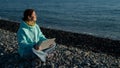 Happy caucasian woman working on a laptop while sitting on a pebble beach.