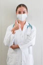 Happy Caucasian young female doctor wearing protective facemask and white hospital gown gives a thumbs up and look at camera, Royalty Free Stock Photo