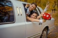 Happy charming newlywed couple is looking through the car window. The bride with the wedding bouquet of red roses is Royalty Free Stock Photo