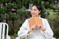 A happy Asian woman sits on a chair with a book in her hand, relaxing in her beautiful green garden Royalty Free Stock Photo