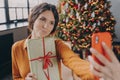Happy charming young brunette woman taking selfie with Christmas gift box on smartphone at home Royalty Free Stock Photo